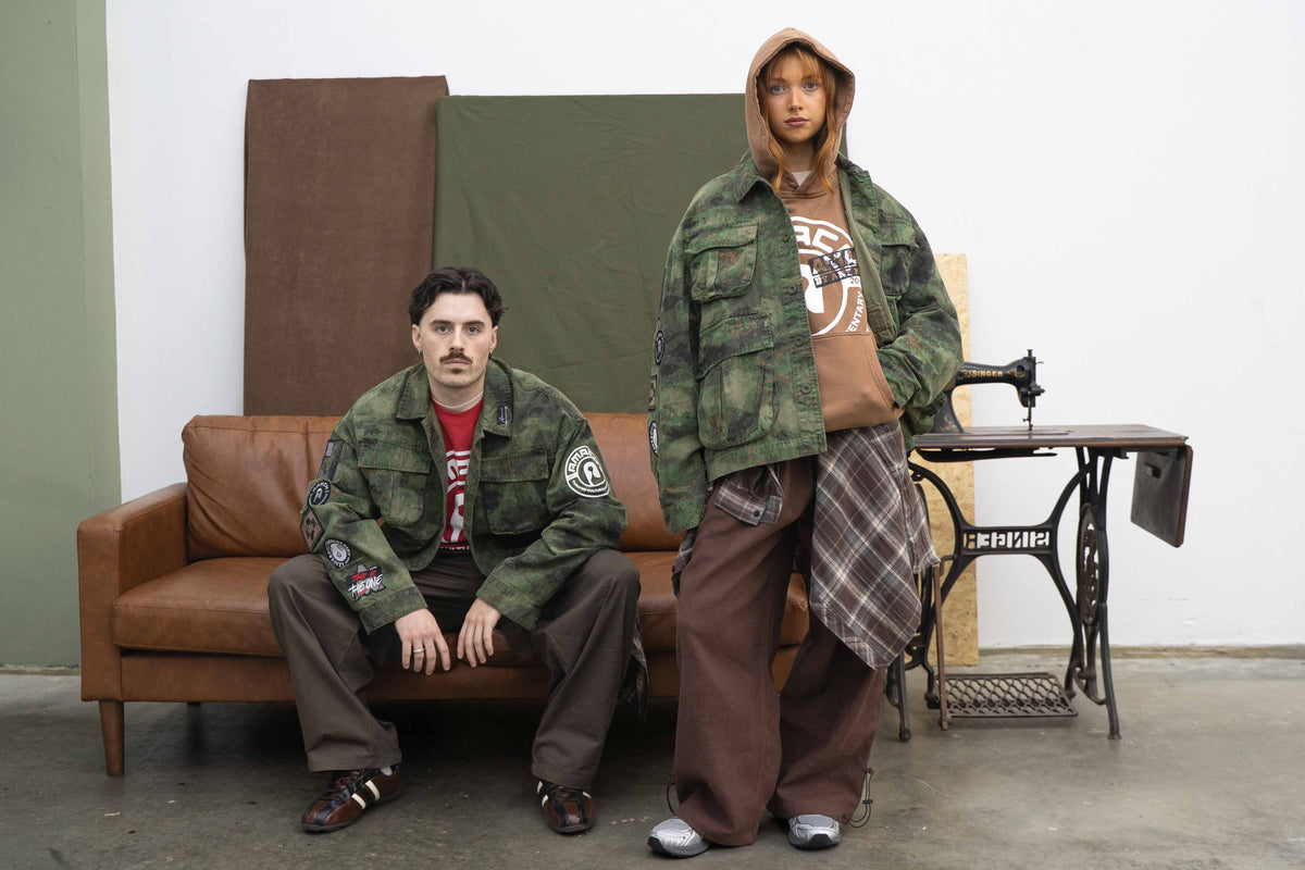 AMACH By Any Means camo military jackets with patches, worn on a leather sofa in the studio