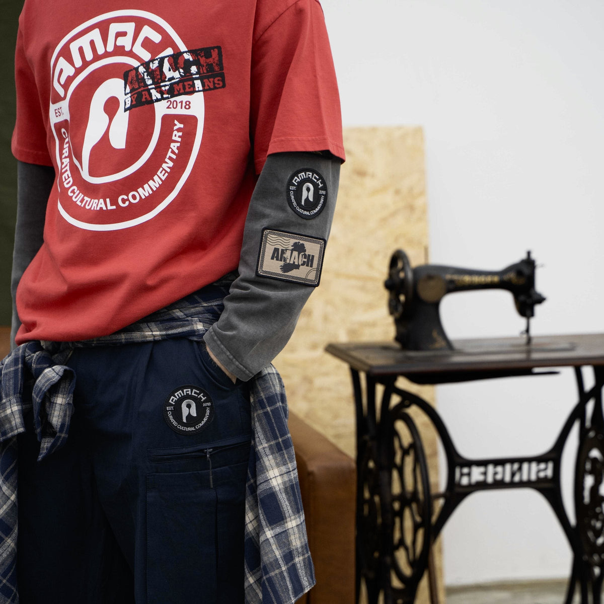AMACH By Any Means red t-shirt layered over a grey long-sleeve, beside a sewing machine